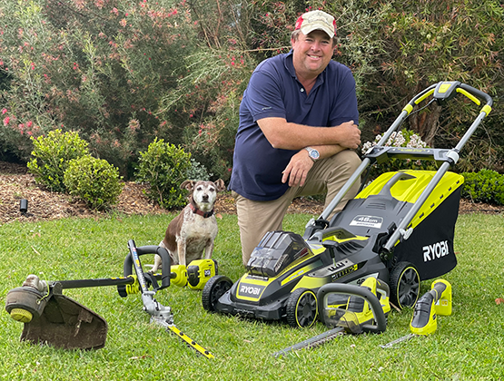 Jason Hodges poses on grass in front RYOBI outdoor products including lawn mower and brushcutter.