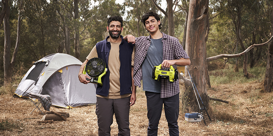 Two men stand, smiling in their camp ground holding RYOBI camping tools